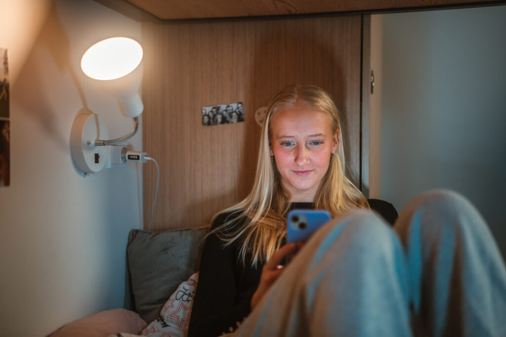 Efterskoleelev tjekker beskeder fra Zenbi på mobilen i ro og mag på sit værelse – digital skolekommunikation med overblik og nærvær.